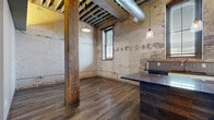 Located at 619 E William St in downtown Wichita, Kansas, this single-floor loft masterfully blends historic industrial character with contemporary finishes. The open living area commands attention with soaring exposed-beam ceilings, original heavy timber columns, and aged brick walls — including a striking arched brick alcove — that speak to the building's rich architectural heritage. Dark hardwood-style flooring grounds the space and complements the raw aesthetic throughout.

The kitchen is a standout feature, outfitted with white cabinetry, dark granite countertops, a central island, stainless steel appliances including a range, microwave, and refrigerator, and a tile backsplash. Pendant lighting and an exposed brick accent wall add warmth and character to the cooking space. Large windows allow natural light to filter in, enhancing the loft's airy atmosphere.

The two bathrooms are finished with modern, textured ceramic tile surrounds wrapping the tub-shower combinations, recessed niche shelving, and wood-look tile flooring — offering a clean contrast to the industrial aesthetic of the main living areas. Two closets and a hallway provide functional storage and circulation throughout. The space also includes an additional unclassified room, adding flexibility for various uses.