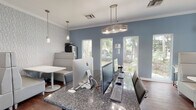 This single-floor lounge in Gainesville, Florida presents a polished, contemporary design that balances functional workspaces with comfortable social seating. The space is characterized by a cool blue-gray color palette on accent walls, complemented by a textured geometric-patterned wallcovering that adds visual depth without overwhelming the room's clean aesthetic.

A central granite-topped workstation serves as the business center hub, equipped with multiple desktop computers and ergonomic seating. The dark, speckled granite surface provides ample workspace and anchors the room with a sophisticated material contrast. Overhead, a multi-tiered oval pendant fixture provides focused illumination above the workstation, while coordinating cylindrical pendant lights hang above the seating area.

The lounge seating area features high-backed upholstered booths in a neutral gray fabric, arranged around white pedestal tables. This layout creates semi-private nooks ideal for small group gatherings or individual use. The geometric-patterned accent wall backdrop reinforces the modern design language throughout.

Dark hardwood flooring runs throughout the space, grounding the lighter furnishings and wall tones. Large windows and glass-paneled doors along one wall invite abundant natural light and offer views of lush outdoor greenery, enhancing the overall brightness and welcoming ambiance of the lounge.