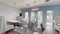 This single-floor lounge in Gainesville, Florida presents a polished, contemporary design that balances functional workspaces with comfortable social seating. The space is characterized by a cool blue-gray color palette on accent walls, complemented by a textured geometric-patterned wallcovering that adds visual depth without overwhelming the room's clean aesthetic.

A central granite-topped workstation serves as the business center hub, equipped with multiple desktop computers and ergonomic seating. The dark, speckled granite surface provides ample workspace and anchors the room with a sophisticated material contrast. Overhead, a multi-tiered oval pendant fixture provides focused illumination above the workstation, while coordinating cylindrical pendant lights hang above the seating area.

The lounge seating area features high-backed upholstered booths in a neutral gray fabric, arranged around white pedestal tables. This layout creates semi-private nooks ideal for small group gatherings or individual use. The geometric-patterned accent wall backdrop reinforces the modern design language throughout.

Dark hardwood flooring runs throughout the space, grounding the lighter furnishings and wall tones. Large windows and glass-paneled doors along one wall invite abundant natural light and offer views of lush outdoor greenery, enhancing the overall brightness and welcoming ambiance of the lounge.
