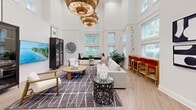 Located at 24080 Amador St in Hayward, California, this thoughtfully designed single-floor space makes a striking impression through its dramatic double-height ceilings that lend an expansive, airy quality well beyond its physical footprint. The living room serves as the centerpiece of the space, showcasing a refined contemporary aesthetic with light hardwood flooring that runs throughout and crisp white walls that amplify the natural brightness.

Towering dual-level windows line the walls, inviting generous amounts of natural light and offering pleasant views of surrounding greenery. A statement cluster of woven rattan pendant chandeliers anchors the room's vertical scale, adding warmth and texture to the lofty ceiling. The seating area features a plush, neutral-toned sofa set atop a patterned area rug, complemented by a round wooden coffee table and an elegant dark glass display cabinet.

A built-in window bench provides an inviting nook for additional seating, while a wall-mounted flat-screen TV and curated artwork contribute to the room's polished character. A high-top bar counter with vibrant red stools adds a functional yet stylish social element to the space. The entryway features a bold chevron-patterned reception desk and vivid wall art, creating a memorable first impression. Overall, the space exudes a bright, modern, and welcoming ambiance.
