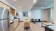 This single-floor unit showcases a contemporary design that blends industrial and modern elements. The open-concept living and kitchen area forms the heart of the space, where light wood-tone flooring runs consistently underfoot. A distinctive architectural detail is the exposed concrete ceiling that transitions into a smooth white-painted surface, lending an urban, loft-like character to the living zone.

The kitchen is efficiently arranged along one wall, featuring light wood cabinetry with open shelving, stainless steel appliances including a gas range, microwave, and dishwasher, and a clean white countertop with an undermount sink. A window above the counter allows natural light to filter in. Adjacent to the kitchen, the dining area accommodates a compact round table, while the living area provides comfortable seating and views through large windows overlooking the surrounding cityscape.

The two bedrooms are simply designed with white walls, light flooring, and windows that bring in natural light. One bedroom includes a built-in desk workspace. Both bathrooms maintain the clean, minimal aesthetic with light wood vanities, white quartz countertops, and full-size tub-shower combinations.