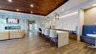 Located at 1401 Kings in Bridgeport, Connecticut, this thoughtfully designed single-floor amenity space blends contemporary aesthetics with functional comfort. The open layout seamlessly connects several distinct zones, each finished with warm wood-look flooring and carefully curated furnishings throughout.

A striking lounge area anchors one end of the space, centered around a dramatic floor-to-ceiling veined marble accent wall housing a linear fireplace and wall-mounted display. Vibrant seating in bold coral and yellow tones adds energy, while large floor-to-ceiling windows flood the area with natural light and provide direct views of an outdoor terrace equipped with seating and grilling amenities.

The kitchen and social hub features a generous quartz-topped island with upholstered bar seating beneath sculptural pendant lights suspended from a rich wood-paneled ceiling. A geometric mosaic tile backsplash, built-in appliances, and warm cabinetry complete the well-appointed bar area. Nearby, a cozy banquette with cobalt blue upholstery offers an intimate dining nook.

A corridor lined with contemporary artwork connects the main gathering spaces to additional seating and work areas, creating a cohesive flow throughout. The overall ambiance is polished, welcoming, and modern.