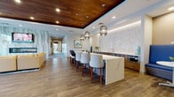 Located at 1401 Kings in Bridgeport, Connecticut, this thoughtfully designed single-floor amenity space blends contemporary aesthetics with functional comfort. The open layout seamlessly connects several distinct zones, each finished with warm wood-look flooring and carefully curated furnishings throughout.

A striking lounge area anchors one end of the space, centered around a dramatic floor-to-ceiling veined marble accent wall housing a linear fireplace and wall-mounted display. Vibrant seating in bold coral and yellow tones adds energy, while large floor-to-ceiling windows flood the area with natural light and provide direct views of an outdoor terrace equipped with seating and grilling amenities.

The kitchen and social hub features a generous quartz-topped island with upholstered bar seating beneath sculptural pendant lights suspended from a rich wood-paneled ceiling. A geometric mosaic tile backsplash, built-in appliances, and warm cabinetry complete the well-appointed bar area. Nearby, a cozy banquette with cobalt blue upholstery offers an intimate dining nook.

A corridor lined with contemporary artwork connects the main gathering spaces to additional seating and work areas, creating a cohesive flow throughout. The overall ambiance is polished, welcoming, and modern.