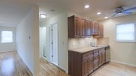 This single-floor space showcases a thoughtfully designed kitchen as a central feature. The kitchen is outfitted with rich, dark-stained wood cabinetry featuring raised-panel detailing and brushed metal hardware, providing generous upper and lower storage. A tile backsplash in neutral tones complements the countertops, while a stainless steel sink with a classic faucet adds a functional focal point. Recessed lighting and a flush-mount ceiling fixture ensure the space is well-lit throughout. Light-toned hardwood flooring runs seamlessly through the kitchen and into the adjacent hallway, contributing warmth and visual continuity. The layout opens to a corridor that leads to additional rooms, including what appears to be a bathroom visible through an open doorway, suggesting a practical and connected floor plan. The overall color palette — warm whites on the walls paired with the medium-toned wood cabinetry and flooring — creates a clean, inviting atmosphere. The space appears well-maintained and move-in ready, with a straightforward, functional design suited to everyday living.