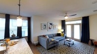 This single-floor residence offers a thoughtfully designed living space that blends comfort and contemporary style. The open-concept living area serves as the heart of the home, anchored by a generous sectional sofa and a bold geometric-patterned area rug that adds visual interest to the wood-style flooring beneath. A ceiling fan overhead provides both function and a finishing touch to the room's clean aesthetic.

Natural light flows abundantly through multiple large windows dressed with dark curtain panels, offering a pleasing contrast against the crisp white walls. A set of glass doors leads to an outdoor area, further enhancing the sense of openness and connectivity to the exterior. Framed artwork adorns the walls, contributing to the curated, modern ambiance of the space.

The living area transitions seamlessly toward a kitchen counter visible at the edge of the frame, suggesting an open-plan layout ideal for entertaining and everyday living. Warm accent lighting from a table lamp complements the natural light, creating an inviting atmosphere throughout the day and into the evening. Overall, the space presents a clean, modern interior with well-considered design details that prioritize both livability and visual appeal.