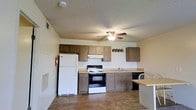 This single-story space is designed with functionality and everyday practicality in mind. The kitchen serves as a central feature, equipped with a full suite of appliances including a white refrigerator, an electric range with oven, and a dishwasher. Medium-toned wood cabinetry lines both the upper and lower walls, providing generous storage space for kitchen essentials. A continuous countertop runs the length of the kitchen, offering ample workspace for meal preparation, and a built-in sink is conveniently positioned along the counter. A ceiling fan with an integrated light fixture is centrally mounted, promoting air circulation and supplemental lighting throughout the space. The flooring features durable tile in a neutral tone that complements the overall color palette of light, neutral walls. A textured ceiling adds subtle visual depth to the room. The layout is open and efficient, with a small dining area adjacent to the kitchen, making the space well-suited for daily living. Overall, the unit presents a straightforward and well-organized interior that prioritizes usability and comfort.
