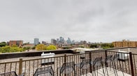 This rooftop space delivers a striking urban experience, highlighted by unobstructed panoramic views of a major downtown skyline. The open-air deck features a gravel surface underfoot, providing both a functional and aesthetically fitting foundation for an elevated outdoor setting. Sturdy metal railings line the perimeter, ensuring safety while preserving the unimpeded sightlines toward the city's towering architecture and surrounding neighborhoods. A collection of metal chairs is thoughtfully arranged across the deck, offering ample seating for guests to take in the expansive cityscape. The rooftop's elevated position places visitors above the surrounding rooftops, creating a sense of openness and scale that is difficult to replicate at ground level. The backdrop of the city skyline — with its mix of high-rise towers, mid-rise buildings, and mature tree canopy — lends the space a dynamic, ever-changing character throughout the day. Whether used for gatherings, events, or quiet reflection, this rooftop terrace provides a compelling and memorable setting defined by its commanding urban views and straightforward, functional design.