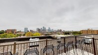 This rooftop space delivers a striking urban experience, highlighted by unobstructed panoramic views of a major downtown skyline. The open-air deck features a gravel surface underfoot, providing both a functional and aesthetically fitting foundation for an elevated outdoor setting. Sturdy metal railings line the perimeter, ensuring safety while preserving the unimpeded sightlines toward the city's towering architecture and surrounding neighborhoods. A collection of metal chairs is thoughtfully arranged across the deck, offering ample seating for guests to take in the expansive cityscape. The rooftop's elevated position places visitors above the surrounding rooftops, creating a sense of openness and scale that is difficult to replicate at ground level. The backdrop of the city skyline — with its mix of high-rise towers, mid-rise buildings, and mature tree canopy — lends the space a dynamic, ever-changing character throughout the day. Whether used for gatherings, events, or quiet reflection, this rooftop terrace provides a compelling and memorable setting defined by its commanding urban views and straightforward, functional design.