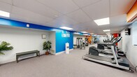 Located at 500 West Rosedale Avenue in West Chester, PA, this fitness center spans a single floor and is divided into two dedicated exercise rooms, offering a well-equipped environment for a comprehensive workout experience. The main exercise area is bright and open, featuring a drop-panel ceiling with recessed lighting that evenly illuminates the space. Bold accent walls in blue and orange energize the environment, with a motivational "No Pain No Gain" mural serving as a focal point. The room is outfitted with a range of cardio equipment, including treadmills, elliptical machines, and stair climbers, all arranged to allow comfortable circulation. Full-length mirrors along one wall amplify the sense of space and allow users to monitor their form. A wall-mounted television provides entertainment during workouts. The secondary exercise room offers a more flexible layout, with storage cubbies, a wall-mounted split AC unit, and a set of double doors with glass panels that invite natural light into the space. This room also provides storage for free weights and additional fitness accessories. Soft carpet flooring throughout both rooms ensures comfort underfoot. The overall ambiance is clean, functional, and motivating, making this facility a practical and well-appointed space for fitness activities.