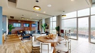 Located at 511 S Mangum St in the heart of Durham, NC, this generously sized rooftop amenity level offers a sophisticated blend of indoor comfort and outdoor living. The interior lounge is anchored by a striking horizontal wood-slat accent wall lined with mounted flat-screen displays, complemented by warm hardwood-style flooring, bold contemporary artwork, and lush tropical greenery. Dramatic floor-to-ceiling windows flood the space with natural light while framing sweeping views of the Durham skyline and the adjacent ballpark below. A built-in wet bar with wood cabinetry, open shelving, and stainless steel appliances provides convenient service for gatherings. Statement pendant lighting with gold-toned fixtures adds a warm, upscale ambiance throughout. The interior flows seamlessly through sliding glass doors onto an expansive outdoor terrace shaded by a modern louvered pergola. Outside, multiple seating arrangements cater to both intimate conversations and larger groups, while a fully equipped outdoor kitchen features professional-grade grills set against a colorful mosaic tile accent wall. Raised planters with ornamental grasses add natural texture, and the open-air terrace offers unobstructed panoramic views of downtown Durham and the surrounding landscape, making it an exceptional space for entertaining and relaxation.