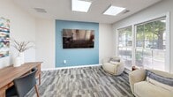 This dedicated fitness studio is a thoughtfully designed exercise space that offers a focused environment for a variety of workout routines. The room features multi-toned patterned carpet tiles throughout, lending a modern, practical aesthetic underfoot. A standout architectural element is the overhead skylight, which floods the space with natural light and contributes to an open, airy atmosphere.

One wall is painted in a calming steel blue accent color, providing visual contrast against the neutral white walls that surround the rest of the room. Mounted on this accent wall is an Echelon Reflect interactive fitness mirror, offering a versatile, tech-forward workout solution. Adjacent to it, a vertical ball storage rack holds multiple exercise balls in assorted sizes and colors, keeping the space organized and accessible.

Large windows along one wall frame a lush outdoor view, further enhancing the bright, welcoming environment. A full-length wall mirror adds to the sense of space and provides functional feedback during workouts. Yoga and exercise mats stored in a woven basket offer convenient access for floor-based training.

The studio connects to an adjacent fitness room visible through glass-paneled doors and interior windows, where cardio equipment including treadmills can be seen. Entry is provided via a glass-paned exterior door, adding to the room's connectivity and natural light. Overall, this studio is a versatile, well-lit fitness space designed for comfort and functionality.