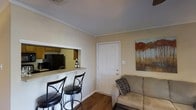 This single-floor space offers a practical and comfortable layout designed to maximize functionality. The living area features warm beige walls, hardwood-style flooring, and crown molding that adds a refined touch throughout. A textured ceiling completes the classic interior aesthetic.

A key highlight is the pass-through breakfast bar that connects the living area to the kitchen, creating an open and interactive layout. The bar is topped with a light-colored counter surface and accommodates two cushioned bar stools with wrought iron frames, making it ideal for casual dining or entertaining. The kitchen, visible through the pass-through, includes dark cabinetry, a black refrigerator, and upper wooden cabinets, offering ample storage.

The living area is furnished with a cushioned sofa accented by a decorative pillow, positioned near the front entry door. A framed landscape painting featuring warm autumn tones adds a decorative focal point to the wall. The front door, finished in white with a six-panel design, provides a welcoming entryway. Overall, the space presents a cozy and efficiently designed interior suited for everyday living.