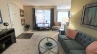 Located at 100 Hunt Club Ln in Raleigh, North Carolina, this single-floor residence offers a well-designed layout that balances comfort and functionality. The living area serves as the heart of the space, featuring plush carpeting underfoot and warm, neutral wall tones that create an inviting atmosphere. Large windows dressed with elegant floor-length drape panels allow an abundance of natural light to fill the room, enhancing the sense of openness and airiness. The layout accommodates a generous seating arrangement, including a sofa with accent pillows and a patterned armchair, all centered around a circular glass-top coffee table that adds a modern touch. A media console along the back wall provides practical storage and display space, while thoughtfully placed floor lamps contribute to the warm, layered lighting of the room. Decorative wall art and potted greenery add character and a lived-in charm to the space. The overall ambiance is calm and comfortable, with a cohesive color palette of soft greens, grays, and warm neutrals that tie the space together seamlessly. This single-level home offers an accessible and easy-flowing floor plan well-suited for everyday living in the heart of Raleigh.