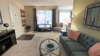 Located at 100 Hunt Club Ln in Raleigh, North Carolina, this single-floor residence offers a well-designed layout that balances comfort and functionality. The living area serves as the heart of the space, featuring plush carpeting underfoot and warm, neutral wall tones that create an inviting atmosphere. Large windows dressed with elegant floor-length drape panels allow an abundance of natural light to fill the room, enhancing the sense of openness and airiness. The layout accommodates a generous seating arrangement, including a sofa with accent pillows and a patterned armchair, all centered around a circular glass-top coffee table that adds a modern touch. A media console along the back wall provides practical storage and display space, while thoughtfully placed floor lamps contribute to the warm, layered lighting of the room. Decorative wall art and potted greenery add character and a lived-in charm to the space. The overall ambiance is calm and comfortable, with a cohesive color palette of soft greens, grays, and warm neutrals that tie the space together seamlessly. This single-level home offers an accessible and easy-flowing floor plan well-suited for everyday living in the heart of Raleigh.
