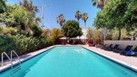 This single-story property showcases a standout outdoor area centered around a large, rectangular swimming pool with vivid turquoise water. The pool is framed by clean, light-colored coping and surrounded by a spacious deck that provides ample room for lounging and relaxation. Tall palm trees and mature deciduous trees create a lush, verdant backdrop, offering natural shade and a sense of seclusion despite the open sky above. Seating areas are thoughtfully arranged around the poolside, including shaded umbrella tables and chairs positioned toward one end, along with sleek chaise lounges along the perimeter — ideal for soaking in the abundant natural sunlight. A distinctive pod-shaped lounge chair adds a stylish, resort-like accent to the space. The surrounding landscaping, featuring dense shrubs and bamboo-style greenery, reinforces a private, tropical ambiance. The overall character of the outdoor space evokes a casual yet inviting retreat, well-suited to warm, sun-drenched climates.