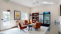 Located at 1710 NE Multnomah St in Portland, Oregon, this single-floor amenity space is designed to serve as a comfortable and fully functional common area. The space is anchored by a bright, open lounge featuring polished terrazzo flooring, recessed lighting, and crisp white walls that lend a clean, modern aesthetic throughout.

The lounge area is furnished with mid-century-inspired leather armchairs and glass-topped tables, creating an inviting spot to relax. Natural light filters in through multiple windows, offering views of the surrounding greenery. A convenience retail corner provides easy access to snacks and beverages, including refrigerated drink units, well-stocked shelving, and a self-service kiosk. A dining counter with tabletop seating rounds out the amenity zone.

Adjacent to the lounge, a dedicated laundry room houses multiple stacked washer and dryer units equipped with digital payment systems, including app-based payment capability. The laundry area features hexagonal tile flooring and a contrasting dark accent wall, adding visual interest to the functional space.

A glass-walled fitness center is accessible from the lounge, offering cardio equipment, ceiling fans, and ample natural light. The seamless flow between the lounge, laundry, and fitness areas makes this a comprehensive, resident-focused amenity hub.