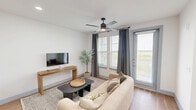 Located at 12820 Langston Blvd in Baytown, TX, this single-story 1-bedroom, 1-bathroom residence offers a well-designed layout that maximizes comfort and functionality. The open-concept living room and kitchen form the heart of the home, with the living area featuring wood-look flooring, a ceiling fan with light, and large windows dressed with gray drape panels and horizontal blinds that invite abundant natural light. The kitchen is a standout, showcasing warm wood-tone cabinetry extending to the ceiling, a large quartz waterfall island with an undermount sink and bar seating, stainless steel appliances, a white tile backsplash, and pendant lighting overhead. The spacious bedroom is carpeted for comfort, features natural light through sizable windows with blinds and curtains, a ceiling fan, and direct access to the dedicated closet and bathroom. The bathroom is finished with a wood-tone vanity, white quartz countertops, a wide mirror with bright overhead lighting, and large-format light tile flooring alongside a walk-in tiled shower. Gray-blue interior doors add a cohesive design detail throughout the home, creating a modern yet warm aesthetic.