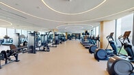 Located at 201 Clearwater Dr in West Palm Beach, FL, this upscale fitness center offers a generously proportioned, single-floor exercise space designed with both function and aesthetics in mind. The room is defined by its standout architectural ceiling detail — large, concentric circular LED light rings that illuminate the space with a warm, ambient glow, creating a polished and energizing atmosphere.

Floor-to-ceiling windows line one side of the facility, flooding the interior with abundant natural light and offering sweeping outdoor views of the West Palm Beach skyline and surrounding landscape. These views are particularly prominent from the cardio zone, where a row of treadmills, stationary bikes, and elliptical machines are positioned to take full advantage of the panoramic scenery.

On the opposite side of the room, full-length mirrors extend across the walls, visually expanding the space and reflecting the extensive lineup of strength training equipment, including cable machines, multi-functional weight stations, free weight racks, and adjustable benches. Warm wood accent panels punctuate the mirrored walls, adding a refined touch to the overall design.

The flooring throughout is a durable, neutral-toned gym surface that complements the bright, open aesthetic. The layout is thoughtfully organized to provide clear separation between cardio and strength training zones, ensuring an efficient and comfortable workout experience.