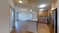 This single-floor space is designed with an open-concept layout that integrates the kitchen and living area into one cohesive environment. The kitchen is a central feature, anchored by a white island with a dark countertop and an undermount sink, providing both functional prep space and a natural dividing point between the kitchen and living area. Warm-toned wood cabinetry lines the walls, complemented by matching dark countertops and ample upper and lower storage. Pendant lights hang above the island, adding focused task lighting with a decorative touch, while flush-mount ceiling fixtures ensure the space remains well-lit throughout.

The flooring is consistent hardwood-style planking that extends from the kitchen through the living area, lending visual continuity and warmth. The living area is open and spacious, offering flexibility for various furniture arrangements. Neutral white walls keep the atmosphere bright and adaptable. A window at the far end of the living space introduces natural light. Visible HVAC vents suggest climate control throughout. Overall, the space presents a clean, functional, and welcoming interior suited for everyday living.