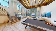 This two-story clubhouse offers a thoughtfully designed community space that blends modern aesthetics with a warm, coastal-inspired atmosphere. The main floor is expansive and open, anchored by dramatic double-height ceilings adorned with rich, dark-stained wood beams and elegant pendant lighting. Large floor-to-ceiling windows flood the space with natural light and provide views of the surrounding exterior. Wood-look tile flooring runs throughout, adding warmth and continuity.

The game room is a central highlight, featuring a full-size pool table and a shuffleboard table, complemented by comfortable lounge seating arranged around a wall-mounted TV set against a striking teal shiplap accent wall. Adjacent to the game area, a fully appointed kitchen offers stainless steel appliances, a large island with bar seating, custom cabinetry, and a decorative tile backsplash — ideal for entertaining.

A dedicated workspace on the main floor includes glass-partitioned private study rooms alongside open desk areas, providing functional and focused environments. A sleek staircase with modern metal railings leads to the second floor, where an additional lounge nook overlooks the main level below. A glass-walled fitness area is also accessible from the main floor, completing this well-rounded and versatile clubhouse facility.