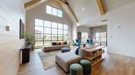 This single-floor space showcases a sophisticated blend of modern design and warm, inviting details throughout. The main lounge area commands immediate attention with its dramatic vaulted ceiling supported by exposed wood beams, paired with expansive floor-to-ceiling black-framed windows that flood the room with natural light and offer sweeping outdoor views. Light hardwood flooring runs throughout, creating a cohesive flow from one area to the next.

The open lounge is thoughtfully arranged with generous sectional seating, accent chairs in rich jewel tones, and layered rugs that define conversation areas. A fireplace set within a sleek dark surround anchors a separate seating vignette, complemented by additional windows that frame outdoor scenery. A small bistro-style table provides an intimate spot nearby.

The kitchen area features a long, white-topped island with bar seating, warm wood cabinetry, and a striking array of pendant lights hanging against another wall of floor-to-ceiling windows. The layout is both functional and visually open.

A dedicated library lounge offers a quieter retreat, featuring built-in shelving finished in deep navy blue, a plush window seat with decorative pillows, and a pair of elegant wingback chairs arranged around a central table — all set against moody, dark-toned walls that create a refined, intimate atmosphere.