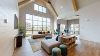 This single-floor space showcases a sophisticated blend of modern design and warm, inviting details throughout. The main lounge area commands immediate attention with its dramatic vaulted ceiling supported by exposed wood beams, paired with expansive floor-to-ceiling black-framed windows that flood the room with natural light and offer sweeping outdoor views. Light hardwood flooring runs throughout, creating a cohesive flow from one area to the next.

The open lounge is thoughtfully arranged with generous sectional seating, accent chairs in rich jewel tones, and layered rugs that define conversation areas. A fireplace set within a sleek dark surround anchors a separate seating vignette, complemented by additional windows that frame outdoor scenery. A small bistro-style table provides an intimate spot nearby.

The kitchen area features a long, white-topped island with bar seating, warm wood cabinetry, and a striking array of pendant lights hanging against another wall of floor-to-ceiling windows. The layout is both functional and visually open.

A dedicated library lounge offers a quieter retreat, featuring built-in shelving finished in deep navy blue, a plush window seat with decorative pillows, and a pair of elegant wingback chairs arranged around a central table — all set against moody, dark-toned walls that create a refined, intimate atmosphere.