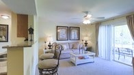 This single-floor residence offers a comfortable and well-appointed living environment designed with a classic, inviting aesthetic. The living room serves as the heart of the space, featuring plush wall-to-wall carpeting that adds warmth underfoot and contributes to the room's cozy atmosphere. Soft, neutral tones on the walls create a calm and welcoming ambiance throughout.

A ceiling fan with integrated lighting fixture is centrally positioned, providing both comfort and illumination. The room is furnished with a light-toned sofa accented with decorative pillows, complemented by a white coffee table and coordinating accent tables, each adorned with elegant table lamps that cast a warm glow. Two framed art pieces with dark borders are symmetrically displayed above the sofa, adding a refined touch to the décor.

Natural light filters into the space through a draped window dressed in flowing curtains, hinting at outdoor access. Bar-style seating near the room's edge suggests a transition to a dining or kitchen area, enhancing the functional flow of the layout. Thoughtful decorative accents, including a birdhouse figurine and coordinating accessories, lend the space a personalized, homey character. Overall, the residence presents a well-balanced blend of comfort, style, and practicality.