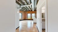 This 2-bedroom, 1-bathroom loft in Syracuse, NY masterfully blends industrial character with modern finishes. The open-concept living and kitchen area serves as the heart of the unit, showcasing soaring exposed timber beam ceilings with visible ductwork, warm hardwood flooring throughout, and original brick walls that add rich texture and historic charm. Large windows flood the space with natural light and frame views of the surrounding cityscape.

The kitchen is well-appointed with crisp white cabinetry, granite countertops, stainless steel appliances including a refrigerator, electric range, and built-in microwave, plus a substantial central island with an undermount sink — ideal for both cooking and entertaining.

The two bedrooms continue the loft's signature aesthetic, with exposed brick accent walls and generous windows offering elevated city views. The primary bedroom provides a comfortable and well-proportioned retreat. The bathroom features clean, neutral tile flooring and bright white walls for a fresh, functional finish.

A dedicated hallway and closet provide practical storage and organization. This unit's industrial-meets-contemporary design makes it a distinctive urban living space in the heart of downtown Syracuse.