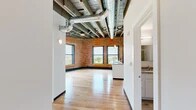 This 2-bedroom, 1-bathroom loft in Syracuse, NY masterfully blends industrial character with modern finishes. The open-concept living and kitchen area serves as the heart of the unit, showcasing soaring exposed timber beam ceilings with visible ductwork, warm hardwood flooring throughout, and original brick walls that add rich texture and historic charm. Large windows flood the space with natural light and frame views of the surrounding cityscape.

The kitchen is well-appointed with crisp white cabinetry, granite countertops, stainless steel appliances including a refrigerator, electric range, and built-in microwave, plus a substantial central island with an undermount sink — ideal for both cooking and entertaining.

The two bedrooms continue the loft's signature aesthetic, with exposed brick accent walls and generous windows offering elevated city views. The primary bedroom provides a comfortable and well-proportioned retreat. The bathroom features clean, neutral tile flooring and bright white walls for a fresh, functional finish.

A dedicated hallway and closet provide practical storage and organization. This unit's industrial-meets-contemporary design makes it a distinctive urban living space in the heart of downtown Syracuse.
