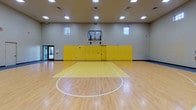 This indoor basketball court is a well-appointed athletic facility designed for serious recreational play. The centerpiece is a full-sized hardwood court finished in a classic light maple tone, with crisp white boundary lines and bold yellow paint marking the key areas, three-point arcs, and center circle. Regulation glass-backboard hoops are mounted at each end, supported by ceiling-suspended assemblies that keep the playing surface unobstructed.

The space rises to notably high ceilings, contributing to a spacious, open atmosphere and providing ample clearance for gameplay. Recessed and flush-panel ceiling lights illuminate the court evenly, supplemented by a row of smaller upper-wall windows that allow additional natural light to filter in. The walls are constructed of painted concrete masonry blocks and are decorated with large-scale mural artwork depicting basketball players in dynamic poses, adding visual energy to the environment.

Along the perimeter walls, yellow wall padding at player-level provides safety near the baselines. Interior windows on multiple walls offer views into adjacent fitness and multipurpose rooms, creating a connected, multi-use facility feel. Entry doors are visible on opposing sides of the court, ensuring convenient access throughout.