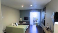This single-floor space offers a thoughtfully designed layout that balances modern aesthetics with everyday functionality. The living area serves as the heart of the space, featuring rich dark hardwood flooring that contrasts beautifully with the light-toned walls and a bold slate-blue accent wall. Recessed lighting in the textured ceiling provides a clean, contemporary look while ensuring the room is well-lit throughout.

A low-profile media console anchors the accent wall, providing a dedicated area for entertainment. Sheer white curtains frame a sliding glass door that opens to a private balcony, flooding the interior with abundant natural light and offering a seamless connection to the outdoors. The balcony can be glimpsed beyond the curtains, hinting at additional outdoor living potential.

The living area is furnished with a soft sage-green sofa accented by decorative pillows, a round side table, and a sleek wire accent chair paired with a glass-top side table — lending the space a curated, contemporary character. A bold typographic wall panel adds a touch of artistic personality. The overall ambiance is modern, light-filled, and inviting, making this space well-suited for both relaxing and entertaining.