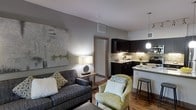This single-floor space showcases a thoughtfully designed open-concept layout that seamlessly connects the living area and kitchen into one cohesive, functional environment. The living area is warm and inviting, anchored by a charcoal-toned sofa and a complementary accent chair in soft green-gold upholstery, creating a balanced and tasteful seating arrangement. A large abstract canvas on the wall adds an artistic focal point, while a classic table lamp provides soft, ambient lighting. Hardwood-style flooring runs throughout, adding warmth and continuity to the space. The kitchen is both stylish and practical, featuring dark espresso cabinetry, a mosaic tile backsplash, and a light-toned countertop that extends into a breakfast bar with bar stool seating — perfect for casual dining. Pendant lighting above the bar area adds a modern touch. The kitchen is well-equipped with full appliances, offering everything needed for everyday cooking. Neutral grey walls and clean white ceilings keep the overall palette fresh and modern, while the open layout promotes an airy, spacious feel. A doorway visible from the living area suggests additional private rooms beyond, completing this well-appointed and contemporary residence.