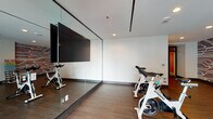 Located at 511 Fayetteville St in downtown Raleigh, NC, this single-floor fitness studio is a well-appointed exercise space designed to support a variety of workout routines. The studio features warm hardwood-style flooring throughout, recessed lighting, and clean white ceilings that create a bright, energizing atmosphere.

A defining visual element of the space is the bold, tech-inspired circuit board mural that spans multiple walls, lending the studio a modern, urban character unique to its downtown Raleigh setting. Floor-to-ceiling mirror panels line one wall, visually expanding the space and providing functional reflection for form-conscious training.

The studio is equipped with professional-grade Spinning bikes positioned in front of a large wall-mounted flat-screen television, making it well-suited for guided cycling classes or individual cardio sessions. A dedicated free-weight area includes a color-coded dumbbell rack alongside aerobic step platforms, supporting strength and cross-training workouts.

Glass partition doors connect the studio to an adjacent fitness area and provide views of what appears to be an outdoor pool deck, introducing natural light and a sense of openness. A built-in wood cabinetry unit near the entrance adds a practical storage element. The overall design balances functionality with contemporary aesthetics, making this a fully equipped and visually engaging fitness environment.