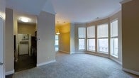 This single-floor space presents a well-designed, open interior with a warm, neutral color palette throughout. The main living area is anchored by a graceful curved bay window configuration, comprising three large windows fitted with white horizontal blinds that allow abundant natural light to filter in while offering views of the surrounding exterior architectural details. Plush wall-to-wall carpeting in a light beige tone extends across the living space, contributing to a comfortable and cohesive aesthetic.

Crown molding runs along the ceiling perimeter, adding a refined architectural detail to the room. An adjacent area, visible through an open passageway, features warm golden-yellow walls and a pendant light fixture, creating a distinct yet connected zone that could serve as a dining or flex space. This secondary area benefits from its own natural light source and continues the carpeted flooring.

To one side, a glimpse into what appears to be a kitchen or utility corridor reveals darker cabinetry and appliances, offering a practical contrast to the lighter living spaces. A visible thermostat on the wall indicates climate control provisions. White baseboards frame each room consistently, tying the overall design together with a clean, polished finish.