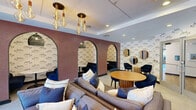 Located at 8 Norden Pl in Norwalk, Connecticut, this thoughtfully designed lounge space offers a compact yet well-appointed environment ideal for relaxed gatherings or casual meetings. The interior is immediately distinguished by its playful nautical-themed wallpaper, featuring illustrated fish motifs that wrap the walls and set a coastal, contemporary tone throughout.

A defining architectural feature is the series of arched alcoves set into a textured, mauve-toned accent wall, each housing intimate seating nooks with small tables and navy blue chairs — perfect for private conversation. The main lounge area is anchored by a plush sectional sofa adorned with layered throw pillows in soft blues and neutrals, complemented by a warm wood round dining table surrounded by coordinating navy chairs.

Overhead, clusters of exposed Edison-style pendant bulbs hang at varying heights, adding a warm, ambient glow to the space. Recessed lighting further enhances the brightness of the room. Whimsical hanging planters along one wall introduce organic greenery, while potted fiddle-leaf fig trees and fresh floral arrangements contribute a lively, welcoming energy.

Large glass entry doors flood the entrance with natural light and provide views of the surrounding outdoor courtyard and neighborhood, seamlessly connecting the interior to its Norwalk setting.