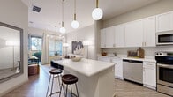 This thoughtfully designed 1-bedroom, 1-bathroom residence offers a modern and cohesive aesthetic across a single floor. The open-concept kitchen and living area serves as the heart of the home, where a large white quartz island with bar seating anchors the space beneath a trio of globe pendant lights with brass accents. The kitchen is outfitted with crisp white shaker cabinets featuring gold hardware, stainless steel appliances including a refrigerator, range, microwave, and dishwasher, and durable wood-look flooring throughout. The living area flows naturally from the kitchen, bathed in natural light from large windows, and opens onto a private balcony through a glass door. The bedroom offers a calm, neutral palette with plush carpeting, natural light from a window with blinds and curtains, and access to a dedicated closet. The full bathroom features a white vanity with gold hardware, a large mirror, tile flooring, and a shower-tub combination. Additional conveniences include a dedicated in-unit laundry area and ample storage. The overall ambiance is contemporary and welcoming, defined by a soft neutral color palette, modern lighting, and clean architectural lines throughout.