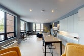 This thoughtfully designed single-floor space functions as a modern communal lounge and gathering area, combining comfort and functionality in a cohesive open-plan layout. The interior is anchored by a bold color palette of deep navy, slate blue, and charcoal gray, complemented by warm gold and mustard accents that add vibrancy throughout.

At the heart of the space, a long communal high-top table with sleek metal legs and a light neutral surface provides ample room for collaborative work or casual dining. Flanking one wall is a fully equipped kitchenette featuring floor-to-ceiling light gray cabinetry, a continuous white countertop, and an integrated sink — offering a clean, streamlined utility zone.

The lounge area is anchored by a plush deep navy sofa adorned with gold and cream throw pillows, positioned to face a wall-mounted flat-screen television. An illuminated built-in shelving unit with backlit display niches serves as both a functional storage solution and a striking focal point, showcasing curated décor and books.

Large black-framed windows line multiple walls, flooding the space with natural light and offering views of surrounding greenery and neighboring architecture. Additional high-top bar seating near the windows creates a flexible zone ideal for casual meetings or individual work. Light wood-toned flooring ties the entire space together, enhancing the bright and inviting atmosphere.