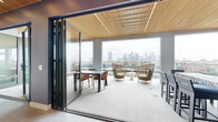 This thoughtfully designed single-floor amenity space combines indoor comfort with outdoor accessibility, highlighted by a striking slatted wood ceiling that flows seamlessly from the interior through to the covered outdoor terrace. Large-format porcelain tile flooring grounds the interior zones, transitioning to a light-toned terrace deck beyond expansive folding glass wall systems that open fully to blur the boundary between inside and out.

The outdoor terrace is a standout feature, furnished with woven rattan lounge chairs, a dining table with colorful seating, and unobstructed panoramic views of a downtown city skyline. Inside, a lounge area offers plush sectional seating, bold yellow accent chairs, a wall-mounted television, and artful decorative lighting against a soft lavender textured wall. Adjacent to it, a game area provides additional entertainment space.

A dedicated bar and dining zone features a striking patterned stone high-top table with bar stools, teal cabinetry, a prep sink, and generous counter space — ideal for social gatherings. An artistic ombre accent wall in shades of teal and dusty rose adds visual depth to the transitional areas of the space, contributing to the overall vibrant yet refined ambiance throughout.