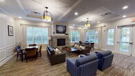 Located at 300 Starboard Side Lane in Rochester, NY, this single-story community space combines classic architectural details with warm, functional design. The main gathering area serves as the heart of the space, anchored by a floor-to-ceiling stone fireplace framed by nautical-themed artwork. Comfortable dark leather sofas and blue striped armchairs are arranged to encourage conversation, while a dining table with wicker and wood chairs provides a more structured seating option. Large windows with patterned drapes flood the room with natural light and offer views of an outdoor deck and surrounding greenery.

A coffered ceiling with recessed lighting and pendant fixtures adds architectural elegance throughout. Crown molding and wainscoting detail the walls in a connected sitting area, which features built-in shelving, a wall-mounted television, and additional seating — ideal for media viewing or quiet reading.

The kitchen is fully equipped with rich dark cabinetry, granite countertops, stainless steel appliances, and ample storage, making it well-suited for group gatherings or catered events. Striped carpeting flows consistently through the common areas, unifying the overall aesthetic. The space presents a polished, community-oriented environment designed for both relaxed socializing and organized activities.