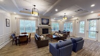 Located at 300 Starboard Side Lane in Rochester, NY, this single-story community space combines classic architectural details with warm, functional design. The main gathering area serves as the heart of the space, anchored by a floor-to-ceiling stone fireplace framed by nautical-themed artwork. Comfortable dark leather sofas and blue striped armchairs are arranged to encourage conversation, while a dining table with wicker and wood chairs provides a more structured seating option. Large windows with patterned drapes flood the room with natural light and offer views of an outdoor deck and surrounding greenery.

A coffered ceiling with recessed lighting and pendant fixtures adds architectural elegance throughout. Crown molding and wainscoting detail the walls in a connected sitting area, which features built-in shelving, a wall-mounted television, and additional seating — ideal for media viewing or quiet reading.

The kitchen is fully equipped with rich dark cabinetry, granite countertops, stainless steel appliances, and ample storage, making it well-suited for group gatherings or catered events. Striped carpeting flows consistently through the common areas, unifying the overall aesthetic. The space presents a polished, community-oriented environment designed for both relaxed socializing and organized activities.