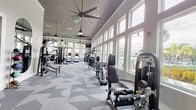 Located at 12231 Pioneers Way in Orlando, FL, this modern fitness center is designed to deliver a comprehensive workout experience across a single, thoughtfully organized floor. The main gym area features soaring vaulted ceilings fitted with large ceiling fans and stylish pendant lighting, creating an airy and energizing atmosphere. Rows of large windows line the walls, flooding the space with natural light and offering views of the surrounding Florida landscape, including swaying palm trees. The patterned geometric carpet throughout the main floor adds a contemporary aesthetic while accommodating a wide array of strength and cardio equipment, including cable machines, weight benches, leg press stations, treadmills, a punching bag, and a squat rack. A dedicated free weights area includes a full dumbbell rack and adjustable bench, complemented by floor-to-ceiling mirrors that enhance the sense of openness. A separate spin studio features wood-look flooring, stationary cycling bikes, a wall-mounted television, arched mirrors, and a bold motivational mural that reads "Underestimate Me. That'll be fun." The studio also includes a rack of color-coded dumbbells and exercise balls for cross-training. Together, these spaces form a dynamic and well-appointed fitness facility built for serious training.