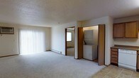 This single-floor space features an open, carpeted living area bathed in natural light from a large window dressed with vertical blinds. The room is finished with a textured ceiling and neutral walls, creating a clean and understated aesthetic. A baseboard heating unit runs along the exterior wall, providing an efficient and low-profile heat source. Adjacent to the main living area, an open doorway leads to a secondary carpeted room, also equipped with a baseboard heater and a window that brings in additional natural light. A dedicated laundry closet with bi-fold doors houses a stacked washer and dryer unit, offering the convenience of in-unit laundry. The flooring transitions from plush carpet in the living spaces to hard-surface tile near the laundry area, providing a practical and durable surface where needed. The overall layout is straightforward and functional, with a neutral color palette throughout that lends itself to easy personalization. The space is well-suited for everyday living, combining practical amenities with a comfortable, low-maintenance interior design.