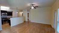 This single-story space showcases an open-concept layout that seamlessly connects the living area to the kitchen. Light hardwood flooring extends throughout, lending warmth and continuity to the interior. The living area is outfitted with a ceiling fan featuring an integrated light fixture, ensuring both comfort and illumination. Neutral, light-toned walls contribute to a bright and airy atmosphere throughout.

The kitchen is partially separated from the living area by a raised breakfast bar counter finished with a granite-style surface, providing both a functional workspace and a casual dining option. Pendant lights hang above the counter, adding a focused decorative touch. A cutout pass-through above the bar maintains an open sightline between the two spaces.

A hallway visible from the main living area leads to additional rooms, with a glimpse of a bathroom beyond. The layout is practical and well-organized, with adequate wall space for flexible furniture arrangements. Ceiling vents are integrated cleanly into the design. Overall, the space presents a clean, functional, and comfortable environment suited for everyday living.