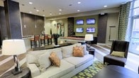 This single-floor lounge is thoughtfully designed for recreation and relaxation, combining modern aesthetics with a range of entertainment amenities. The space is anchored by a plush, neutral-toned sofa adorned with decorative accent pillows, positioned atop a geometric patterned area rug in muted greens and browns. A cluster of four flat-screen televisions mounted on an olive-green feature wall serves as a central focal point, creating an immersive viewing experience. Adjacent to the seating area, a sleek billiards table adds a recreational dimension to the space. Recessed ceiling lighting throughout ensures even, ambient illumination, complemented by a contemporary table lamp that adds warmth. A bar-height counter with upholstered stools runs along one side, offering additional seating and a social gathering spot. Decorative wall panels with rich wood-toned textures and a large mirror enhance the sense of depth and dimension. The overall design palette — warm greens, browns, and creams — creates a cohesive, inviting atmosphere well-suited for both casual gatherings and leisurely downtime.