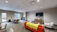 This single-floor clubroom offers a thoughtfully designed communal space that balances comfort and functionality. The interior lounge area features a drop tile ceiling with recessed lighting and patterned carpet flooring throughout. A bold orange sofa anchors the seating area, complemented by neutral accent chairs, a round wooden coffee table, and a wall-mounted flat-screen television above a decorative herringbone-patterned credenza. Abstract artwork adds a touch of color to the neutral gray walls. Adjacent to the lounge, a dining area provides additional seating with a table and chairs, while a small beverage station with dark cabinetry offers convenience for gatherings. A sliding glass door connects the interior to the outdoor courtyard. The courtyard, enclosed by the building's modern metal panel exterior, features large-format tile paving, wicker lounge and dining furniture with orange cushions, string lighting, and a gas grill — making it a versatile outdoor entertaining space. Warm-toned wood slatted privacy screens add character to the exterior setting, creating a welcoming and private atmosphere for residents to gather and relax.