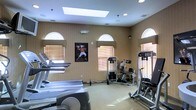 This single-floor fitness center is designed to support a well-rounded workout experience. The room is generously sized, accommodating a variety of cardio equipment—including treadmills and an elliptical machine—alongside a selection of strength training and cable machines. The layout allows for comfortable movement between stations without feeling cramped.

The space benefits from abundant natural light, courtesy of several arched transom windows positioned high along the walls, complemented by a large skylight overhead. Together, these features create a bright, open atmosphere. Neutral-toned, textured wall coverings add a warm yet clean aesthetic throughout.

Recessed lighting and wall-mounted sconces supplement the natural light, ensuring the space remains well-lit at all hours. The flooring appears to be a light-toned, durable surface suited for gym use. Motivational sports artwork adorns one of the walls, contributing to the energizing ambiance. The combination of practical equipment, thoughtful lighting, and airy architectural details makes this fitness center a functional and inviting space for daily exercise.