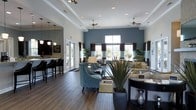 This well-appointed single-floor space showcases a thoughtfully designed open-concept layout that seamlessly blends comfort and contemporary style. Rich, dark hardwood flooring runs throughout, providing a warm foundation that contrasts beautifully with the crisp white tray ceilings adorned with ceiling fans and recessed lighting. A striking deep teal accent wall serves as a bold focal point, adding depth and character to the main gathering area.

The spacious lounge area is furnished with elegant teal accent chairs and neutral-toned sofas, arranged to encourage both conversation and relaxation. Tall, large-pane windows dressed with dark curtains flood the interior with natural light while offering views to the outdoors. Lush potted plants and carefully selected décor add a vibrant, welcoming touch throughout the space.

Adjacent to the main lounge is a bar or café-style area, distinguished by pendant lighting and high seating, ideal for casual gatherings or informal meetings. The transition between zones is smooth and intuitive, maintaining a cohesive design language throughout. Crown molding and tray ceiling details lend an upscale, polished feel, while the overall layout balances openness with defined areas of purpose, making this a versatile and inviting communal environment.