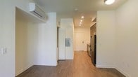This well-appointed single-floor space offers a clean, modern aesthetic with an efficient and functional layout. The open living area features warm hardwood-style flooring that flows seamlessly throughout, creating a cohesive and inviting atmosphere. Crisp white walls and recessed ceiling lighting brighten the interior, contributing to an airy, contemporary feel.

A wall-mounted mini-split HVAC unit provides personalized climate control, while a louvered closet door adds practical storage within the living space. An electrical panel is conveniently accessible along one of the interior walls.

The kitchen is positioned toward the rear of the unit and is equipped with stainless steel appliances, including a full-size refrigerator and range, complemented by rich wood-toned cabinetry. The kitchen's warm finishes offer a subtle contrast to the bright white walls, adding visual depth and character to the space.

A white entry door with a modern hardware finish anchors the end of the hallway, providing a clear and welcoming sense of arrival. The overall design strikes a balance between contemporary minimalism and functional livability, making the space well-suited for comfortable everyday use.