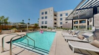 Located on West Sunset Boulevard in the heart of Los Angeles, this amenity level offers a well-rounded collection of shared spaces designed for both relaxation and activity. The centerpiece is a resort-style outdoor pool framed by a spacious sun deck, where rows of chaise lounges and shaded seating areas invite residents to unwind beneath the Southern California sky. Surrounding the pool, lush planted beds and sculptural landscaping soften the clean lines of the contemporary multi-story building, while an outdoor lounge courtyard features built-in seating and a linear fire feature ideal for socializing. A gated pool entry and covered pergola add both security and architectural character to the outdoor environment. Indoors, a bright and airy fitness center is outfitted with cardio machines, a cable strength system, free weights, and exercise balls, benefiting from large windows and direct outdoor access that flood the space with natural light. A stylish interior lounge area features warm wood paneling, large windows overlooking the courtyard, and a modern sectional seating arrangement that creates an inviting gathering space. Adjacent to it, a light-filled kitchen and dining area with light wood cabinetry, white quartz countertops, and large-format tile flooring opens seamlessly to an outdoor terrace with bar seating, blending indoor and outdoor living effortlessly.