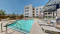Located on West Sunset Boulevard in the heart of Los Angeles, this amenity level offers a well-rounded collection of shared spaces designed for both relaxation and activity. The centerpiece is a resort-style outdoor pool framed by a spacious sun deck, where rows of chaise lounges and shaded seating areas invite residents to unwind beneath the Southern California sky. Surrounding the pool, lush planted beds and sculptural landscaping soften the clean lines of the contemporary multi-story building, while an outdoor lounge courtyard features built-in seating and a linear fire feature ideal for socializing. A gated pool entry and covered pergola add both security and architectural character to the outdoor environment. Indoors, a bright and airy fitness center is outfitted with cardio machines, a cable strength system, free weights, and exercise balls, benefiting from large windows and direct outdoor access that flood the space with natural light. A stylish interior lounge area features warm wood paneling, large windows overlooking the courtyard, and a modern sectional seating arrangement that creates an inviting gathering space. Adjacent to it, a light-filled kitchen and dining area with light wood cabinetry, white quartz countertops, and large-format tile flooring opens seamlessly to an outdoor terrace with bar seating, blending indoor and outdoor living effortlessly.
